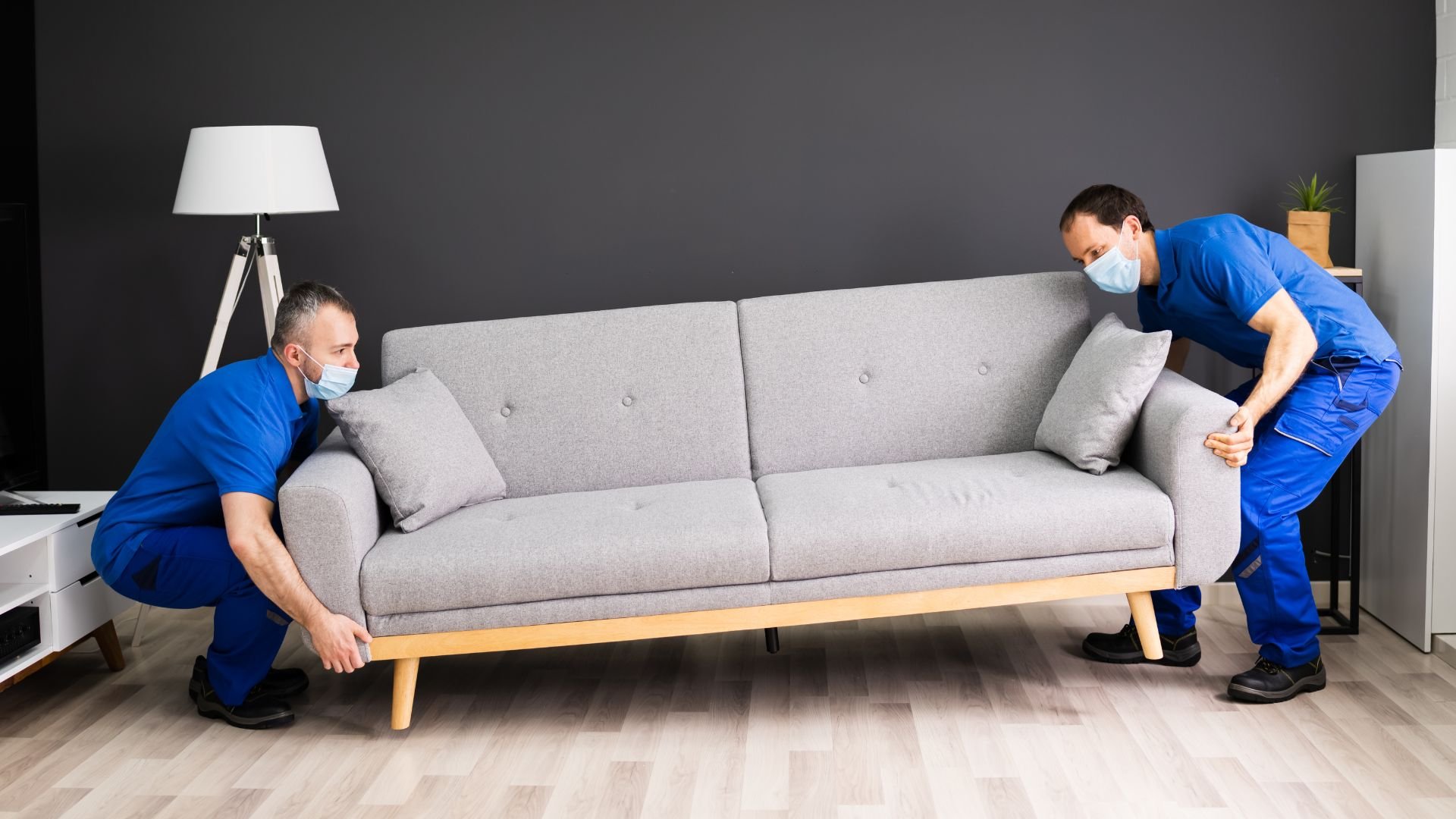 Two movers in blue uniforms lifting a gray couch in a living room