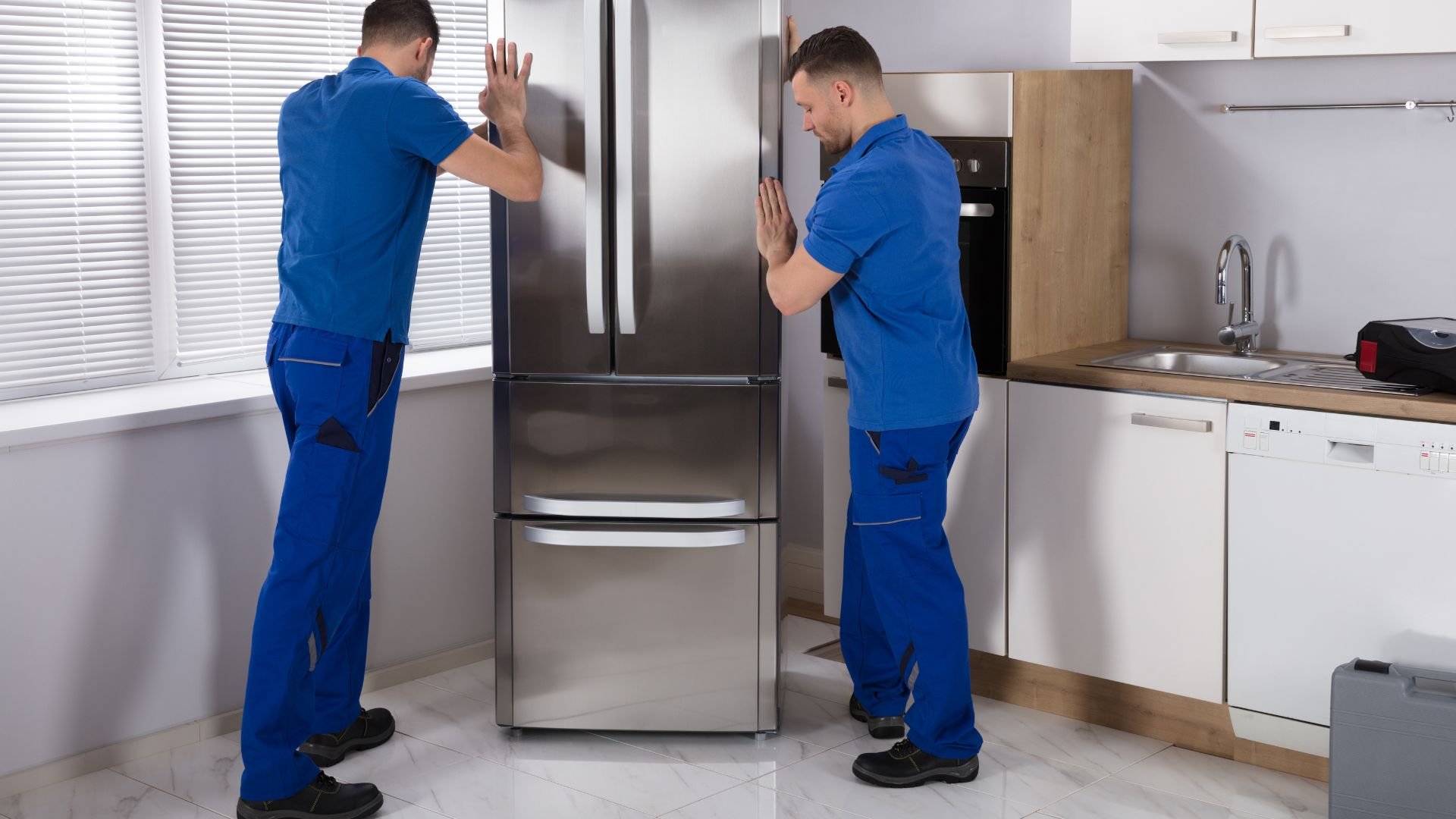 Two technicians in blue uniforms repairing a large stainless steel refrigerator